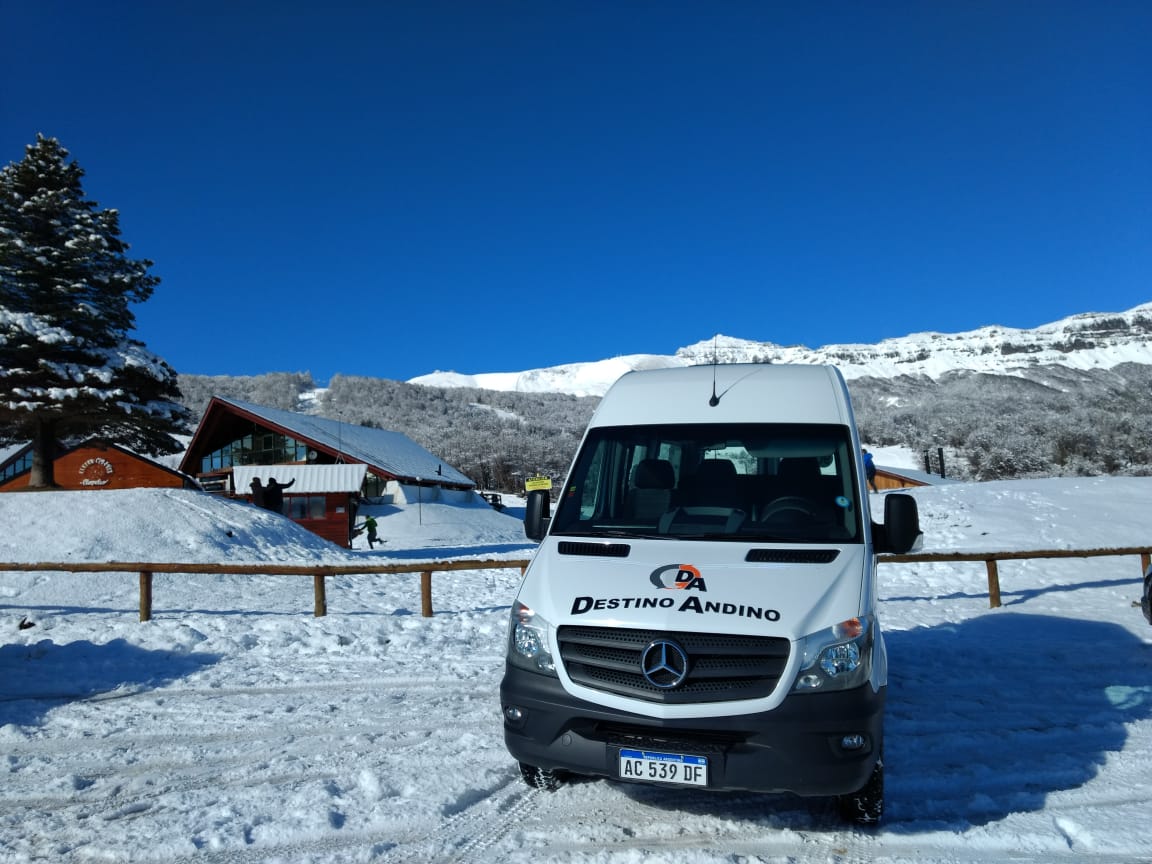 La imagen muestra una van de color blanco, con el logotipo de "DESTINO ANDINO" en el capó. Está estacionada en un paisaje nevado, con mucha nieve cubriendo el suelo y las montañas al fondo. El cielo es de un azul intenso y despejado, lo que sugiere un día soleado de invierno. El sentimiento que genera la imagen es de aventura, majestuosidad y frescura. Invita a pensar en actividades invernales como el esquí o el snowboard, en viajes a la montaña y en la belleza imponente de la naturaleza en invierno. La van de "DESTINO ANDINO" refuerza la idea de un viaje.
