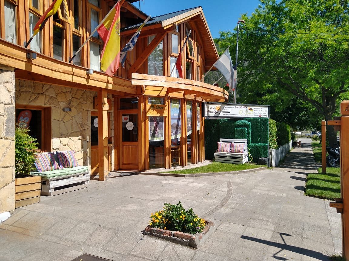 
La imagen muestra la fachada de  una agencia de turismo, con un estilo arquitectónico rústico y acogedor, predominando la madera y la piedra. Hay varias ventanas grandes que reflejan el cielo y la vegetación circundante. En la entrada, se observan banderas de distintas nacionalidades. En el exterior hay bancos de madera con cojines, indicando un espacio para sentarse y relajarse. También hay plantas y un pequeño jardín, lo que contribuye a un ambiente que transmite calma, tranquilidad y bienvenida.