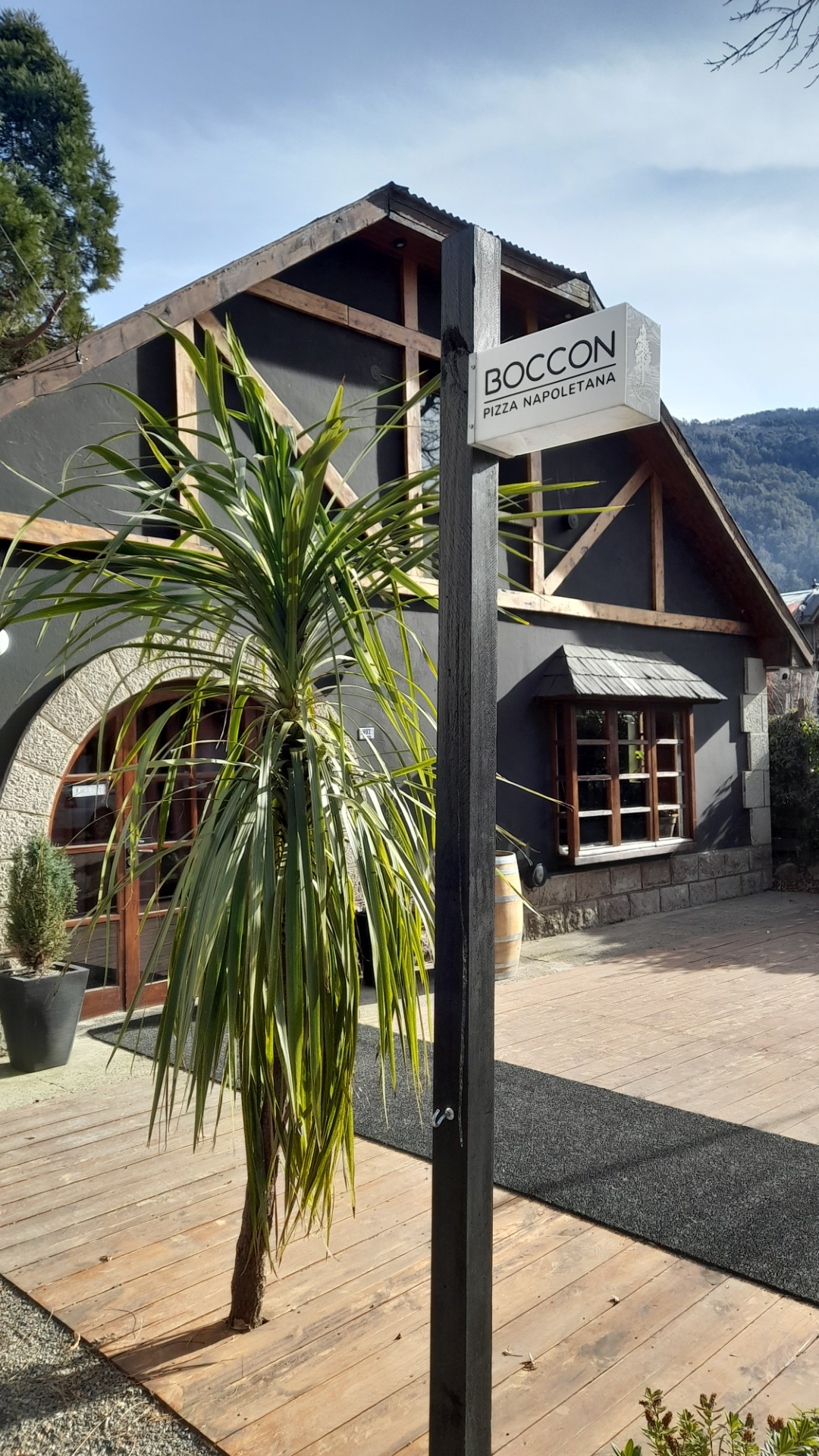 Boccon Pizza Napoletana - Casa de 2 plantas con paredes color gris oscuro, detalles en madera y techo a dos aguas, en la vereda nay plantas y un poste de madera con un cartel rectangular que dice Boccon Pizza Napoletana.