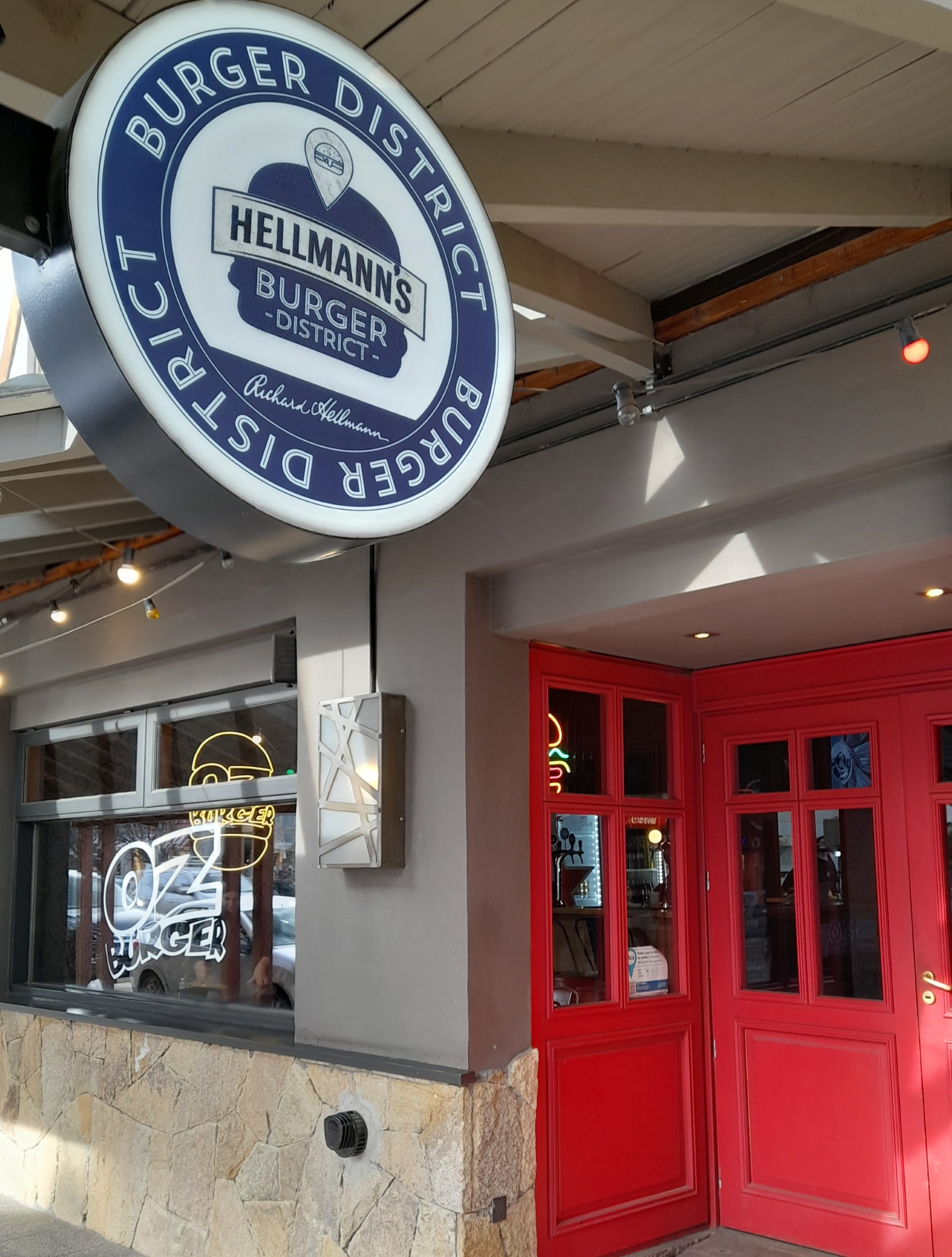 OZ Burger - Fachada de ingreso al restaurante OZ Wine Restó, paredes de piedra, gran ventanal con maderas pintadas en color oscuro y puerta en color rojo. Un cartel colgado en la entrada redondo de fondo blanco y letras azules que dice Burger district.