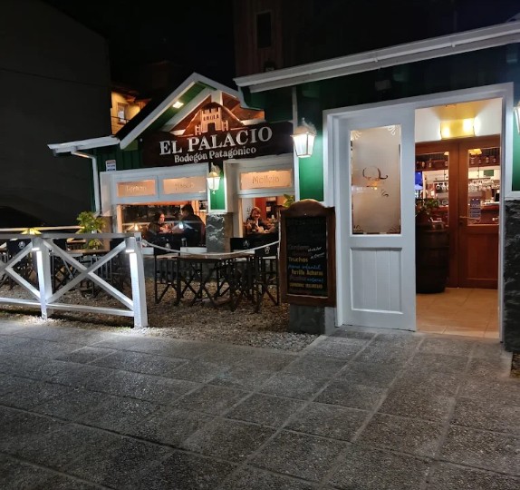 El Palacio- Bodegón Patagónico  - Edificio de paredes de madera pintadas de blanco y verde  con puertas y ventanas  blancas vidriadas, techo a dos aguas. 