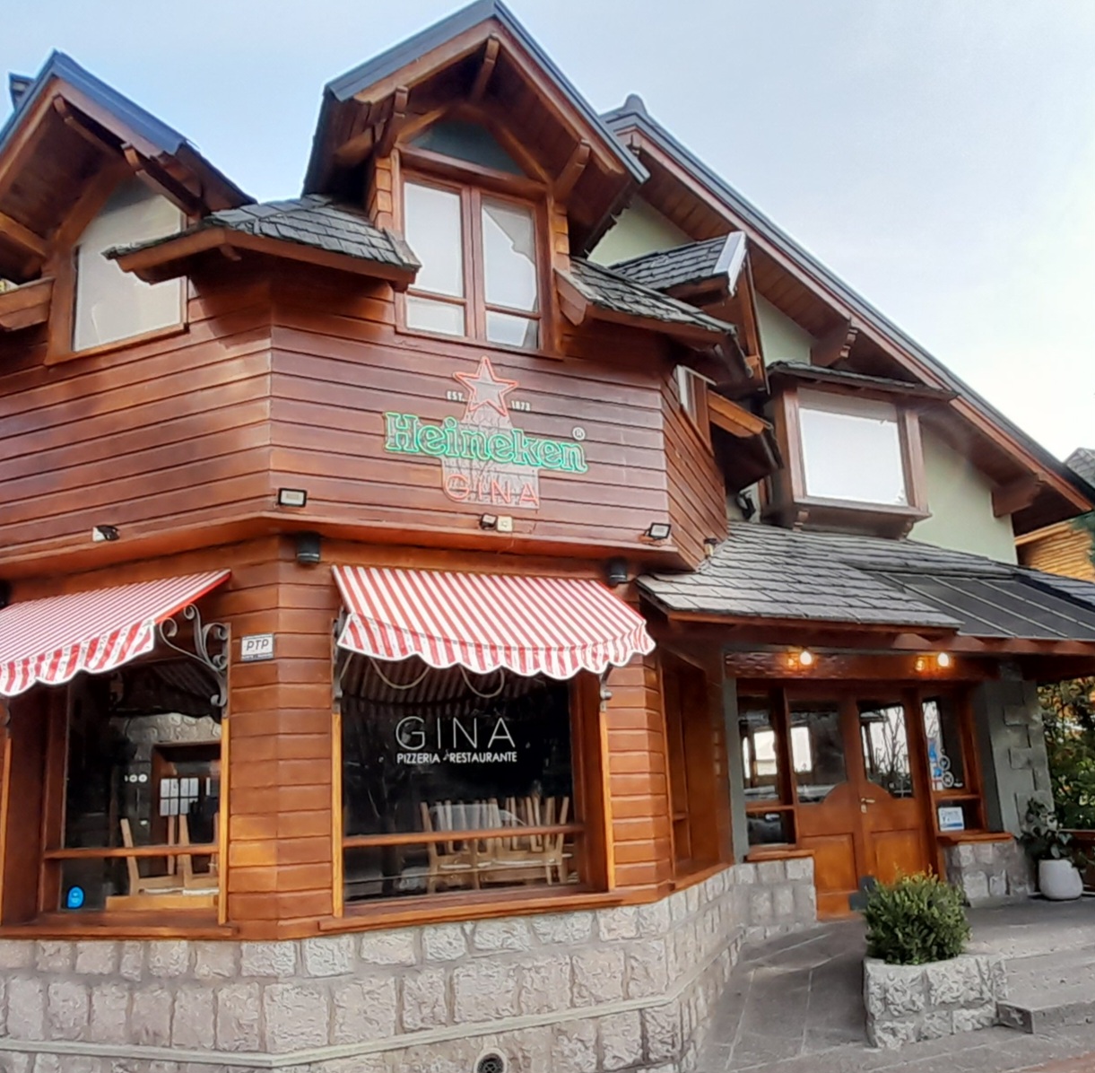 Gina - Entrada al restaurante y pizzería Gina, con puerta de madera y vidrio, parte de las paredes revestidas en madera y el resto de cemento pintada de verde.