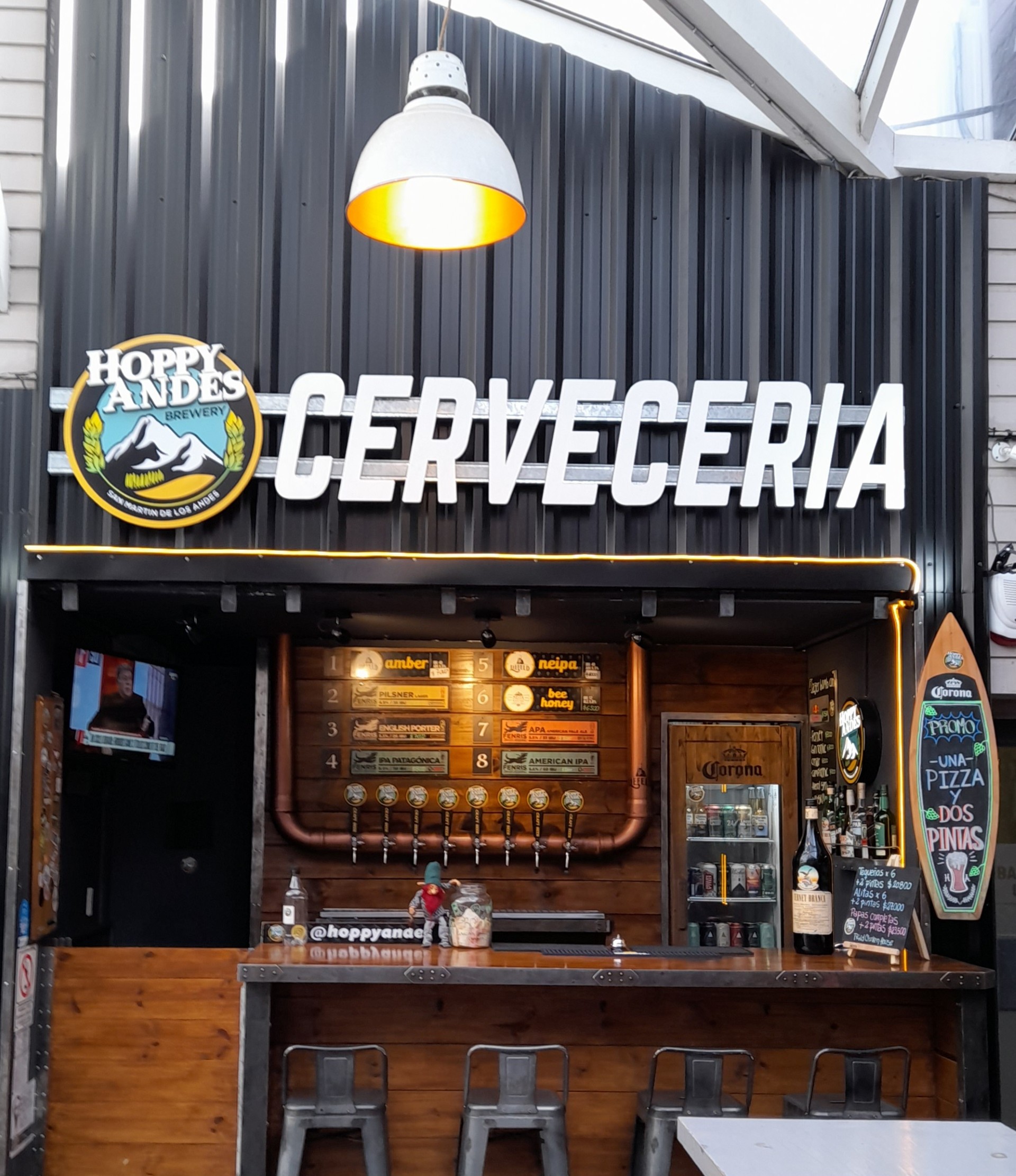 Cervecería Hoppy Andes - La imagen muestra el frente de una cervecería llamada "Hoppy Andes Brewery". Sobre el mostrador principal, un gran letrero luminoso dice "CERVECERIA" en letras blancas. A la izquierda de este, se encuentra el logotipo de la cervecería: un emblema redondo con el nombre "HOPPY ANDES BREWERY" y una imagen de montañas.
mostrador de madera rústica con taburetes frente a él , Detrás del mostrador, se observa un sistema de grifos de cerveza con ocho tiradores, También hay un letrero vertical en forma de tabla de surf que anuncia "UNA PIZZA Y DOS PINTAS". El fondo de la cervecería está revestido con paneles metálicos de color oscuro, lo que le da un aspecto moderno e industrial. Una lámpara colgante blanca con un interior dorado ilumina el área del mostrador desde arriba. En general, la imagen transmite un ambiente acogedor y listo para servir cerveza.