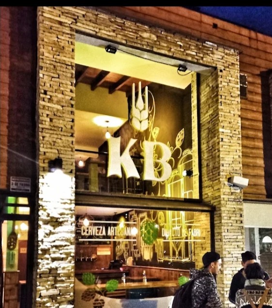 Kraft Brewers Point - Foto nocturna de la fachada de un comercio con dos grandes ventanales que permite ver el interior de una cervecería muy iluminada, en los vidrios tiene dos letras bien visibles que dicen KB, siglas de la cervecería Kraft Brewers, en los costados de las ventanas y en la parte superiores las paredes están recubiertas de piedra y sectores con madera.