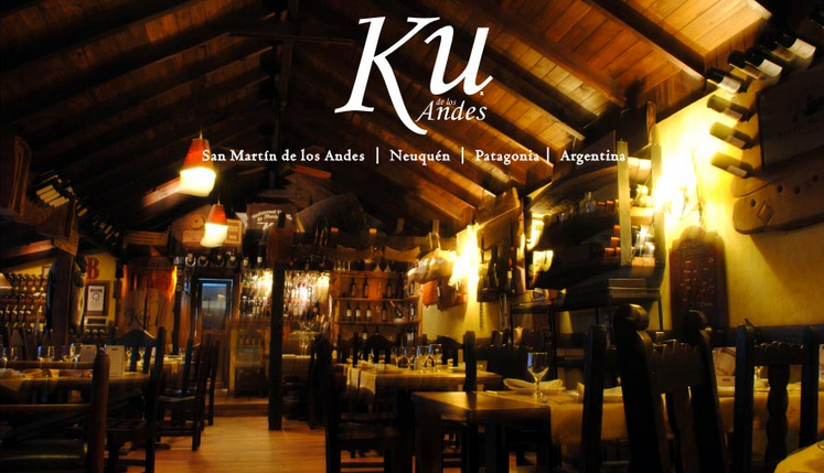 Restaurante Ku - Interior de restaurante con luz tenue, se ven mesas, sillas, techos y pisos de madera, paredes con vitrinas que contienen distinta variedad de vinos.
