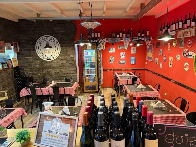 Lo de Chacho trattoria - La imagen muestra un espacio con mesas y sillas, algunas botellas en primer plano, paredes de color con un cartel que indica el nombre del local.