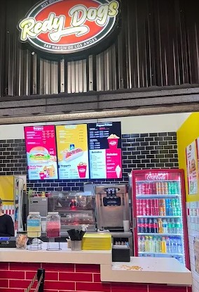 Redy Dogs - En la fotografía hay mostrador de un local de comida rápida. Arriba, ves un letrero luminoso que dice "Redy Dogs", y abajo, la frase "San Martín de los Andes". El fondo es de paneles metálicos oscuros.
El  mostrador es  bajo de ladrillos rojos y blancos. Sobre él, se exhiben bebidas de colores vibrantes en jarras grandes. Detrás del mostrador, en la pared, hay tres pantallas grandes y brillantes con el menú, mostrando fotos de hot dogs, hamburguesas, papas fritas y postres, con sus precios. A la derecha, resalta una nevera vertical roja de bebidas gaseosas llena de latas y botellas de varios colores, brillando intensamente. Se siente como un lugar moderno y lleno de opciones para pedir comida rápida y bebidas refrescantes.