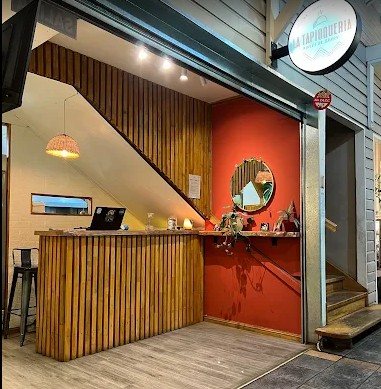 La tapioquería - Comercio que se encuentra dentro de una galería, llamado la tapioquería como está escrito en un cartel redondo con fondo blanco y letras en colores pastel, mostrador y piso en madera, una pared roja con un espejo redondo en el centro, una pared beige y otra con maderas verticales. Sobre el mostrador una lampara que cuelga del techo realizada en mimbre.