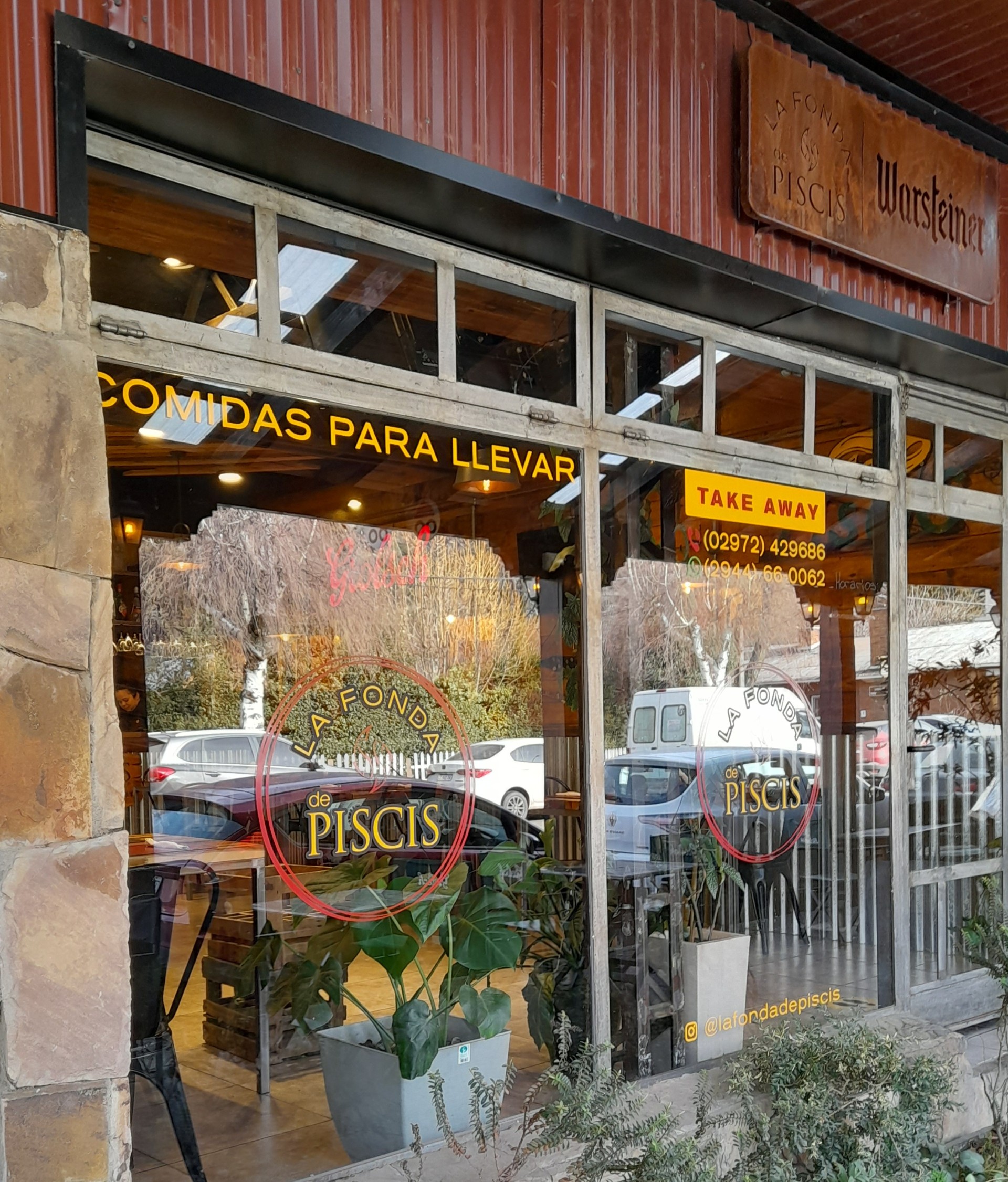 La Fonda de Piscis - Fachada de restaurante y rotisería llamada la Fonda de piscis, tiene grandes ventanales donde tiene el logo del comercio, los marcos de las ventanas son de hierro en color gris, la parte superior decorada con chapa color bordó y las paredes a ambos lados de las ventanas cubiertas con piedras.