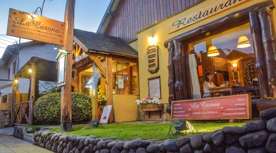 La Casona - Restaurante con techo a dos aguas y paredes de cemento color amarillo con una parte de madera, cartel con el nombre La Casona y jardín en el frente.