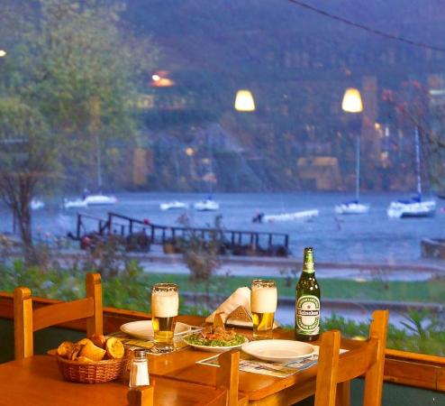 La Costa del Pueblo - Imágen de una mesa con sillas de madera en el interior de un restaurante, al lado de un enorme ventanal desde donde se ve el lago Lácar.