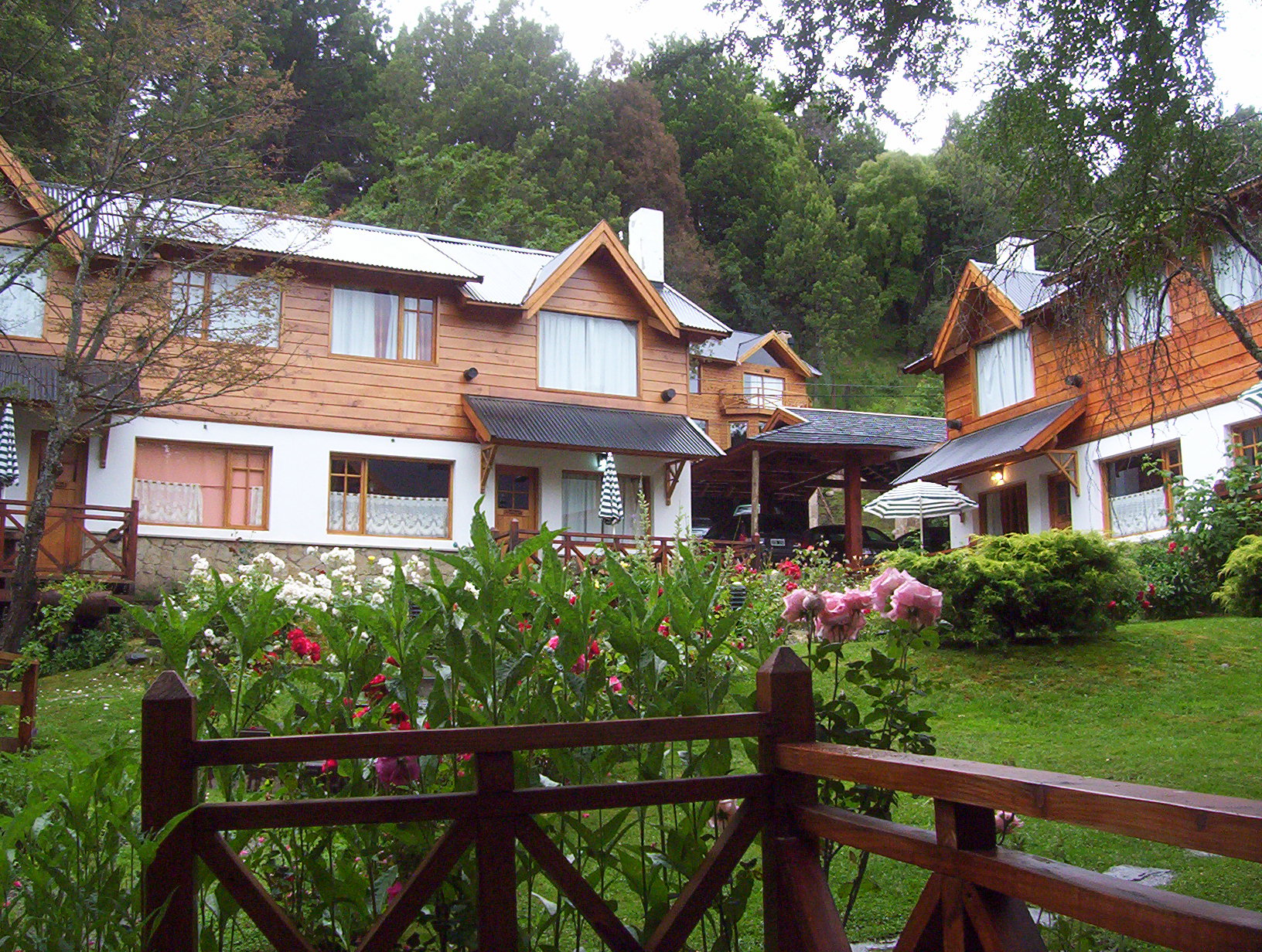 En la imagen se observa un complejo de alojamiento con varias cabañas de estilo arquitectónico de montaña, construidos con una combinación de madera clara y paredes de color claro, con detalles de piedra en la base. Los techos son a dos aguas y las ventanas son amplias, permitiendo la entrada de luz natural. El entorno es muy verde, con césped bien cuidado y abundantes jardines llenos de flores. la imagen transmite tranquilidad, calidez y conexión con la naturaleza.