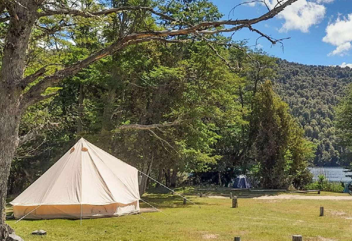 La imagen muestra un glamping  en un camping junto a un bosque y un cuerpo de agua, bajo un cielo azul y soleado.