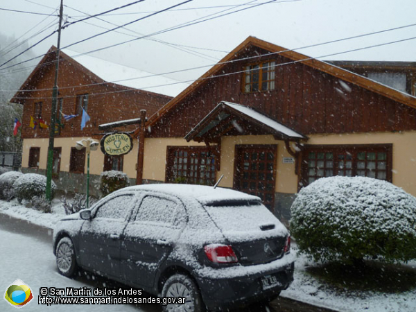 Edificio sencillo, con techo a dos aguas, construido en dos plantas con detalles en la parte superior de madera, al igual que las ventanas y puertas.  La imagen es invernal con nieve en la vereda y techos de la hostería. 