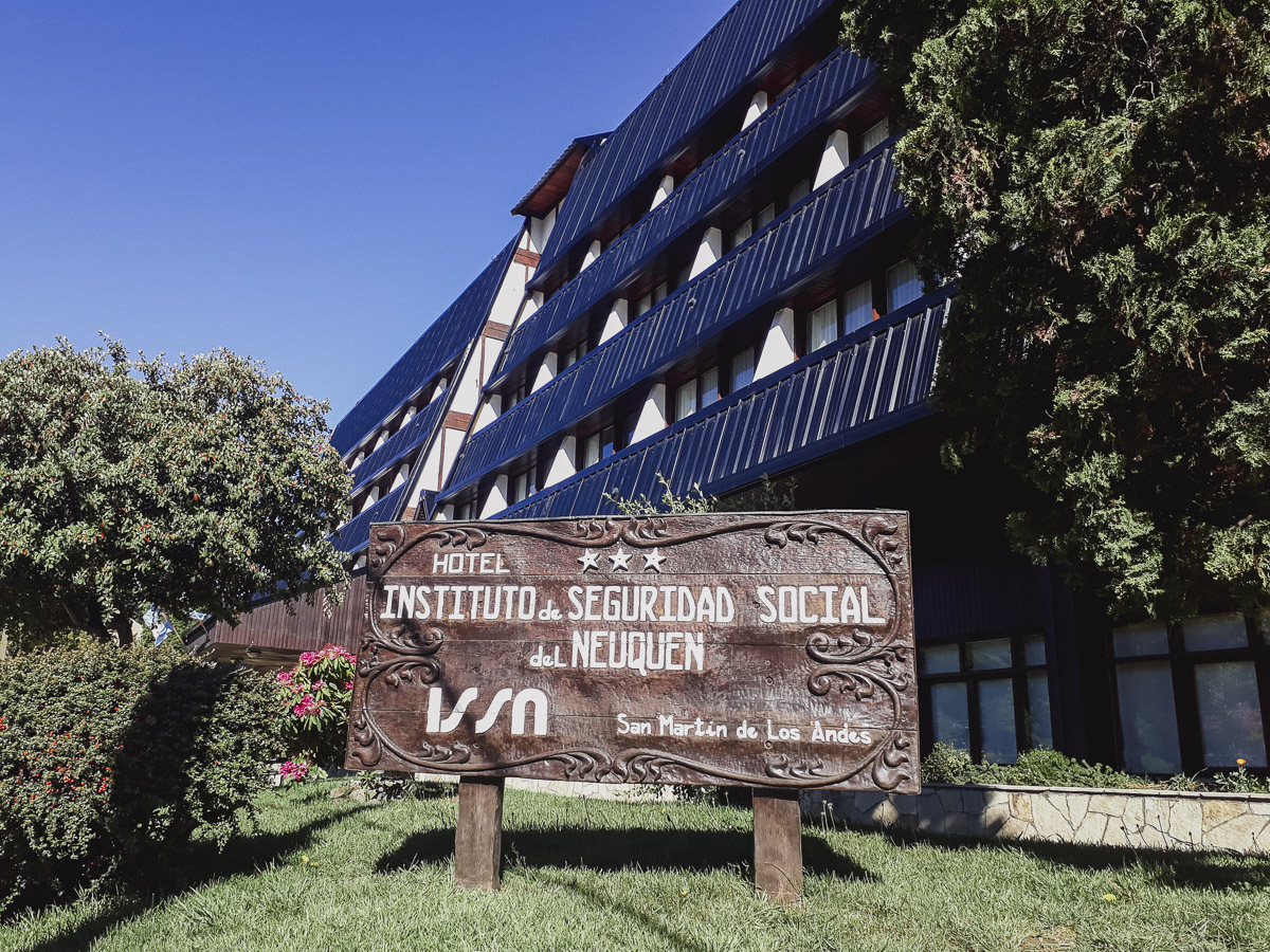La imagen muestra la fachada de un alojamiento de varios pisos con un diseño arquitectónico moderno y distintivo, ubicado en un entorno natural con montañas visibles en el fondo. Tiene una forma piramidal escalonada, con múltiples pisos y ventanas rectangulares. En la parte inferior se ve vegetación bien cuidada, como arbustos y árboles pequeños, y el hall de entrada al hotel.
La estructura del edificio transmite una sensación de solidez y un carácter más contemporáneo que otras construcciones de estilo alpino tradicionales. Puede evocar la idea de un servicio de calidad, funcional y con todas las comodidades de un establecimiento grande, ideal para un viaje de negocios o una estancia confortable.