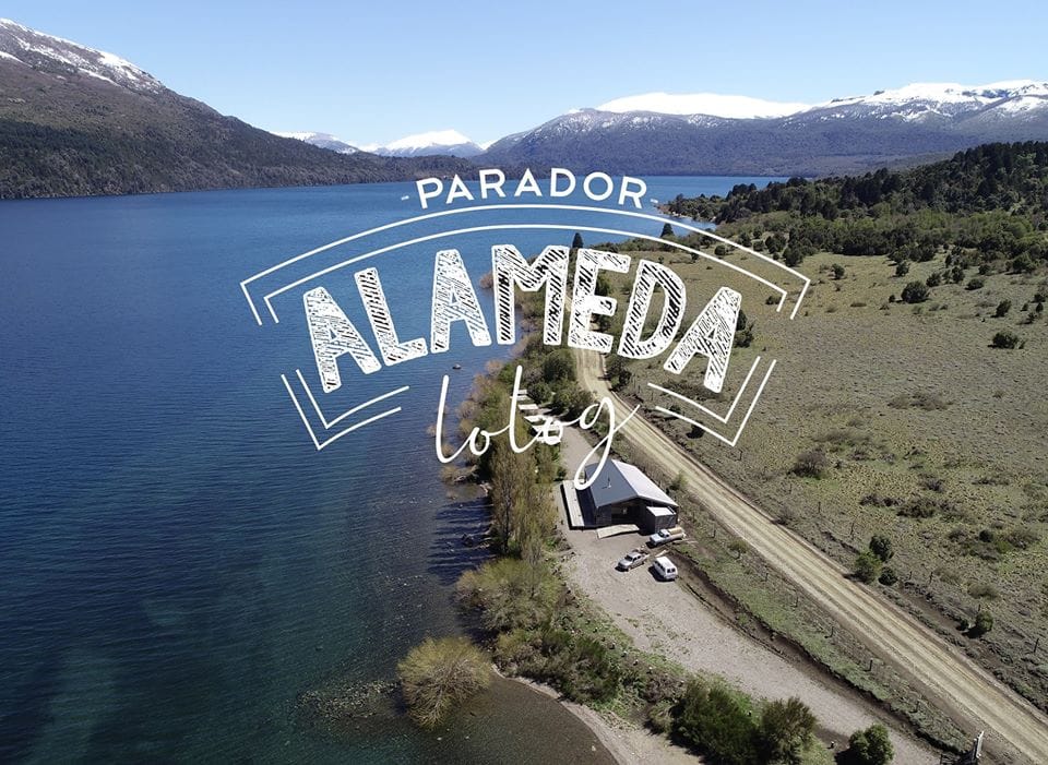 Vista aérea donde se ve el lago Lolog con sus aguas azules, la montaña de fondo y una ruta bordeando el lago. Entre la ruta y el lago el parador La Alameda