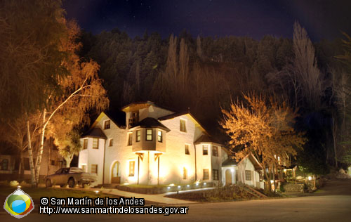 La imagen muestra la hostería La Raclette, edificio de dos pisos con un diseño arquitectónico distintivo, iluminado desde el interior en una noche oscura. Tiene paredes blancas y techos a dos aguas pronunciados. Se pueden ver montañas oscuras en el fondo. Los árboles iluminados lo rodean, creando un ambiente mágico. La imagen transmite una fuerte sensación de cuento de hadas y misterio debido a la iluminación nocturna y el estilo arquitectónico. Evoca calidez y un ambiente acogedor.