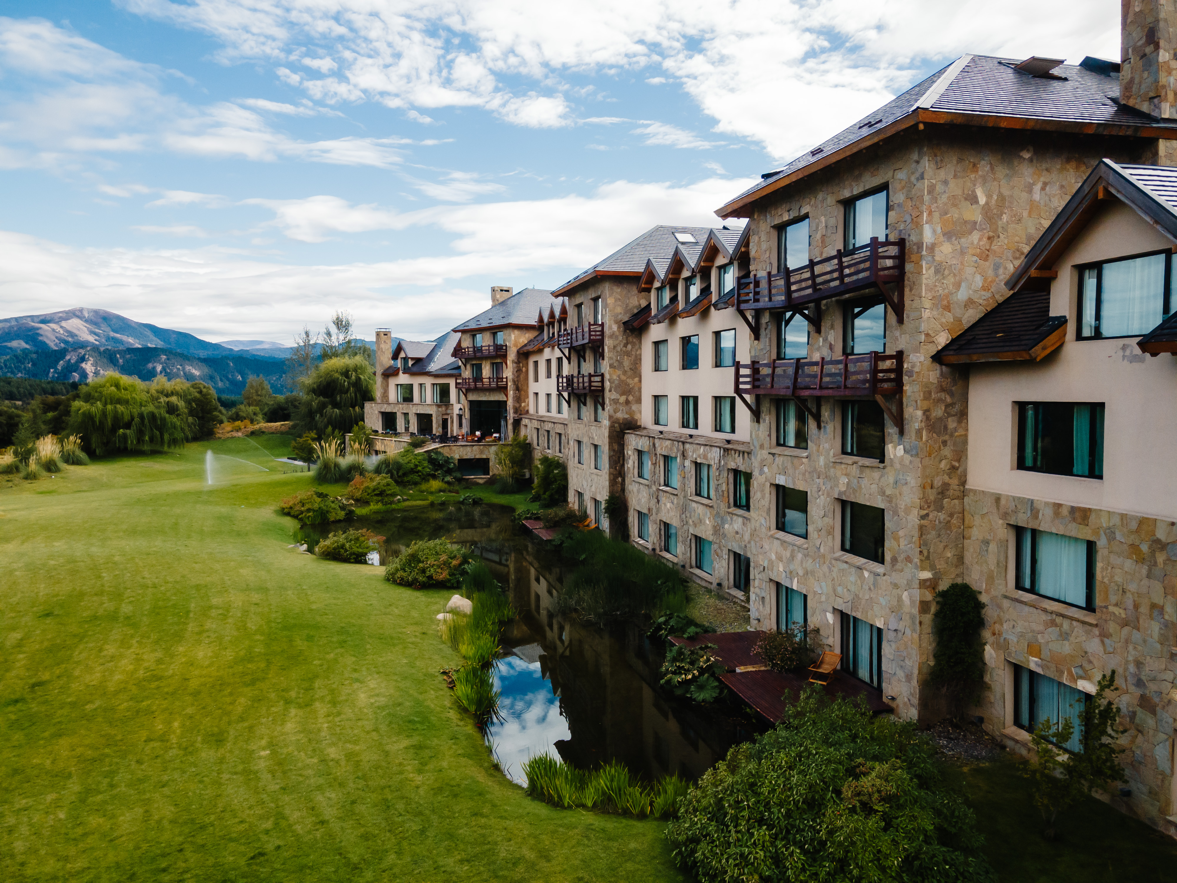 La imagen muestra un gran edificio de estilo rústico elegante, construido con piedra y madera, con varios pisos y balcones del resort de lujo. A la izquierda del edificio, hay un extenso campo de césped muy bien cuidado, parte de un campo de golf (se aprecia una bandera blanca indicando un hoyo). También se observa un pequeño curso de arroyo que corre paralelo al edificio y al césped, con algunos arbustos y vegetación bien mantenida a sus orillas. la imagen evoca una sensación de tranquilidad, lujo y serenidad. La combinación del edificio imponente con el paisaje natural bien cuidado (césped, agua, montañas) sugiere un lugar de descanso, relajación y disfrute de la naturaleza.