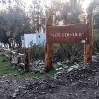 La imagen muestra un cartel de madera con el nombre camping los Coihues,  detrás del cartel se ve el bosque