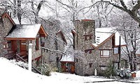 En la imagen se observa un paisaje invernal con varias edificaciones construidas predominantemente en piedra y madera, cubiertas por una capa de nieve. En el centro, destaca una  torre con una arquitectura imponente, hecha de piedra rústica y con techos a dos aguas cubiertos de nieve. A su izquierda, se ve otra cabaña de troncos de madera con techo nevado, y a la derecha, una tercera edificación de piedra con un estilo similar a la central, el suelo está completamente cubierto de nieve, y los árboles del fondo, desprovistos de hojas, la imagen es de serenidad, majestuosidad y un cierto aire de cuento de hadas o fortaleza. La combinación de la piedra y la nieve le da un aspecto robusto y a la vez pintoresco, evocando una sensación de refugio y calidez 
