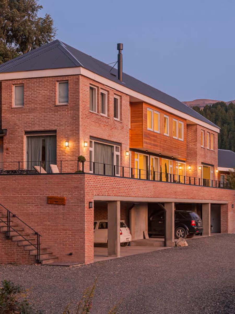En la imagen se observa un edificio construido con ladrillo o mampostería en tonos rojizos o terracota. Tiene varios niveles, con balcones o terrazas que se extienden a lo largo de la fachada y grandes ventanales, algunos de ellos iluminados con una luz cálida, lo que sugiere que es el atardecer o el anochecer. En la parte inferior, se ve un área techada  con arcos utilizados como cocheras, la sensación que da al verlo es de solidez, elegancia y confort. El edificio transmite una imagen de calidad y buen gusto. 