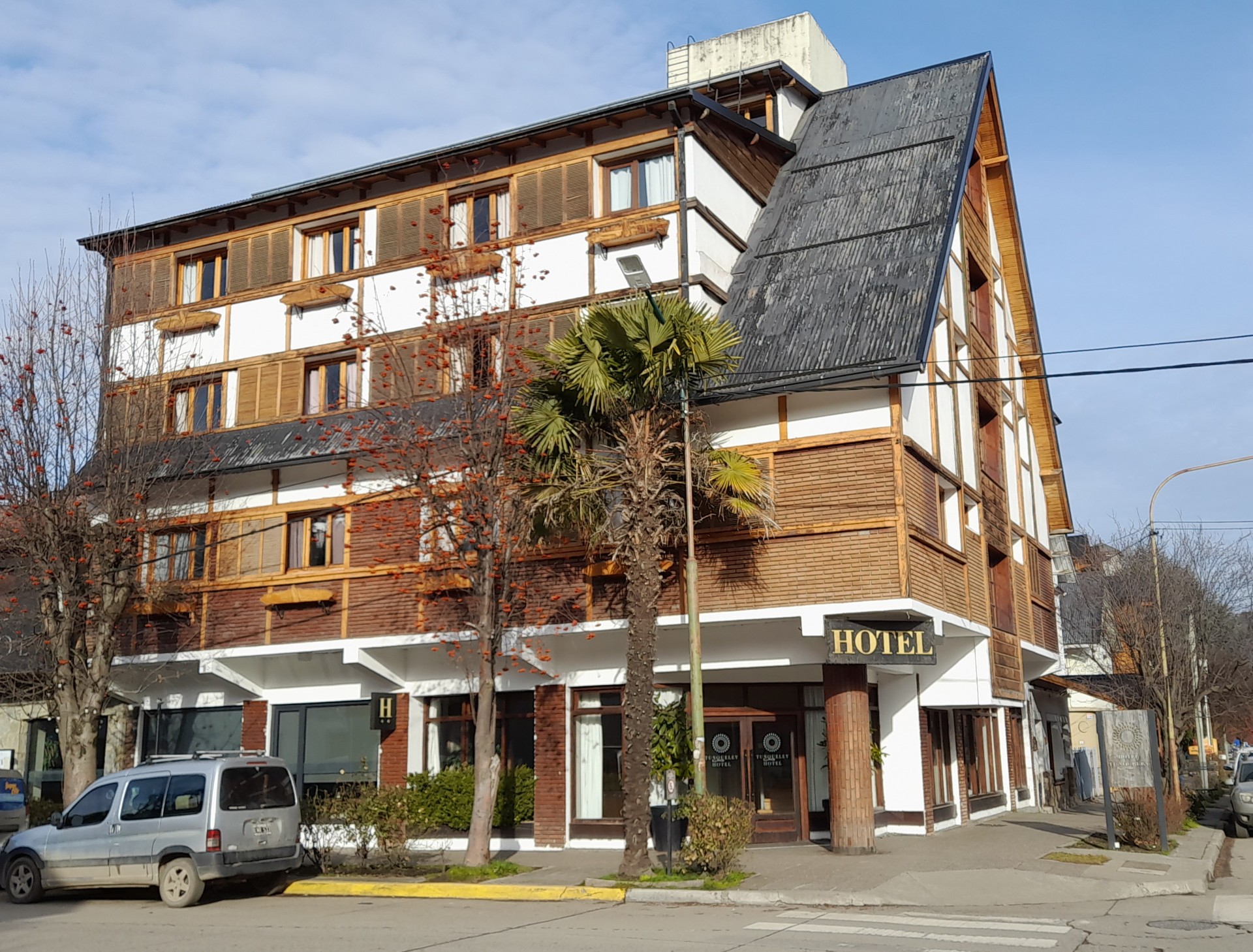 En la imagen se observa un edificio grande de varios pisos, con un estilo arquitectónico que recuerda a las construcciones alpinas o de estilo chalet, con estructura de madera a la vista y paredes en tonos claros y rojizos/marrones. El techo es a dos aguas y pronunciado. En la planta baja, se aprecian escaparates y puertas, La arquitectura imponente y bien cuidada transmite una sensación de calidad y confort. Puede evocar la idea de una estancia agradable en un destino de montaña