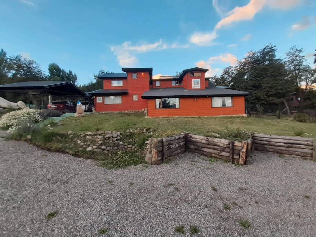 En la imagen se observa una casa de estilo moderno, de color rojo o terracota, con detalles oscuros en el techo y algunas ventanas. Está ubicada en un entorno natural, con árboles y vegetación a su alrededor. El cielo es azul con algunas nubes, lo que sugiere un día despejado.
Al ver la imagen se siente serenidad, modernidad y conexión con la naturaleza. La casa, con su diseño contemporáneo, se integra en el paisaje, transmitiendo una sensación de calma y tranquilidad, evoca la idea de un refugio cómodo y estilizado en un entorno natural.