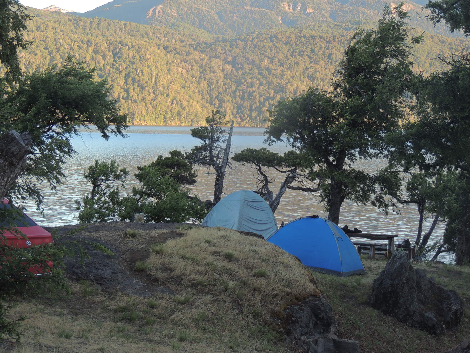 La imagen muestra una zona de acampada rústica en la orilla de un lago.
En el centro se ven dos carpas de campamento. Están montadas en un terreno irregular y con algo de vegetación baja.
El fondo de la imagen dominado por el lago lácar y del otro lado del lago se elevan montañas.
El ambiente general es de naturaleza salvaje y aislamiento, ideal para quienes buscan una experiencia de campamento en un entorno natural.
