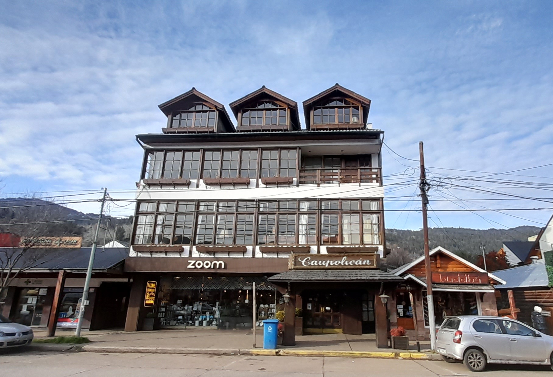 En la imagen se observa el edificio del hotel Caupolican de varias plantas, con un estilo arquitectónico que evoca las construcciones de montaña o centroeuropeas, muy común en San Martín de los Andes. Se destaca por sus balcones de madera en cada piso, sus ventanas con marcos de madera y buhardillas en el techo a dos aguas. La fachada es de color claro, contrastando con los tonos cálidos de la madera.
El edificio transmite una imagen de solidez y un estilo clásico que se integra bien en el paisaje de montaña. Invita a la idea de una estancia cómoda y agradable, ideal para disfrutar de la tranquilidad