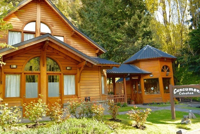 La imagen muestra una  cabaña de estilo alpino, construida principalmente de madera, con techo a dos aguas y ventanas arqueadas, ubicada en un entorno natural con árboles altos y vegetación.