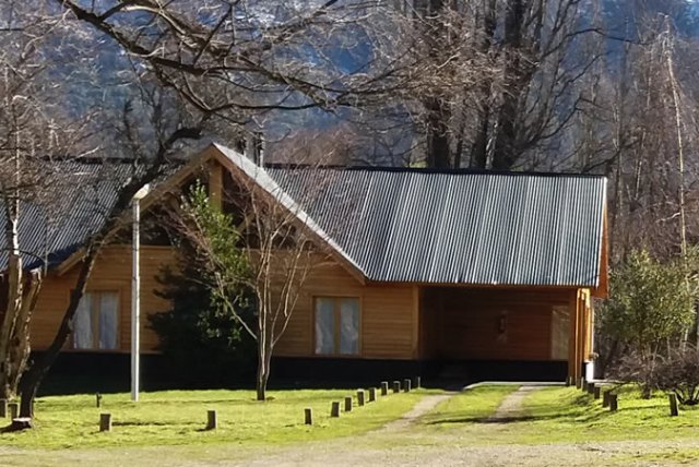 Cabaña de madera de estilo rústico ubicada en un entorno natural rodeada de árboles altos y delgados. La casa, de color marrón claro, presenta un techo a dos aguas distintivo y las ventanas son de color blanco .
