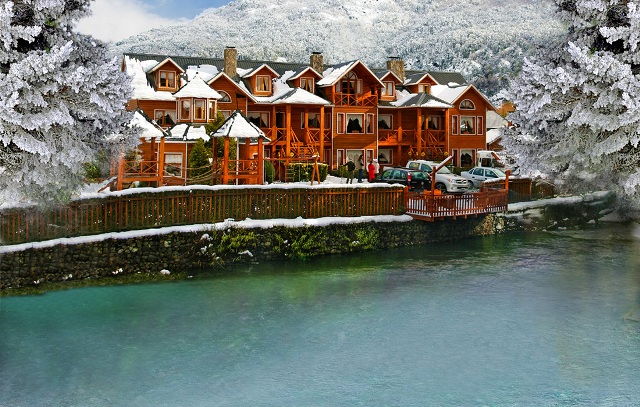 La imagen muestra un edificio de estilo alpino con techo a dos aguas, ubicado a orillas de un arroyo. El edificio está construido principalmente de madera y tiene múltiples ventanas. El paisaje circundante está cubierto de nieve, lo que sugiere que la foto fue tomada durante el invierno.