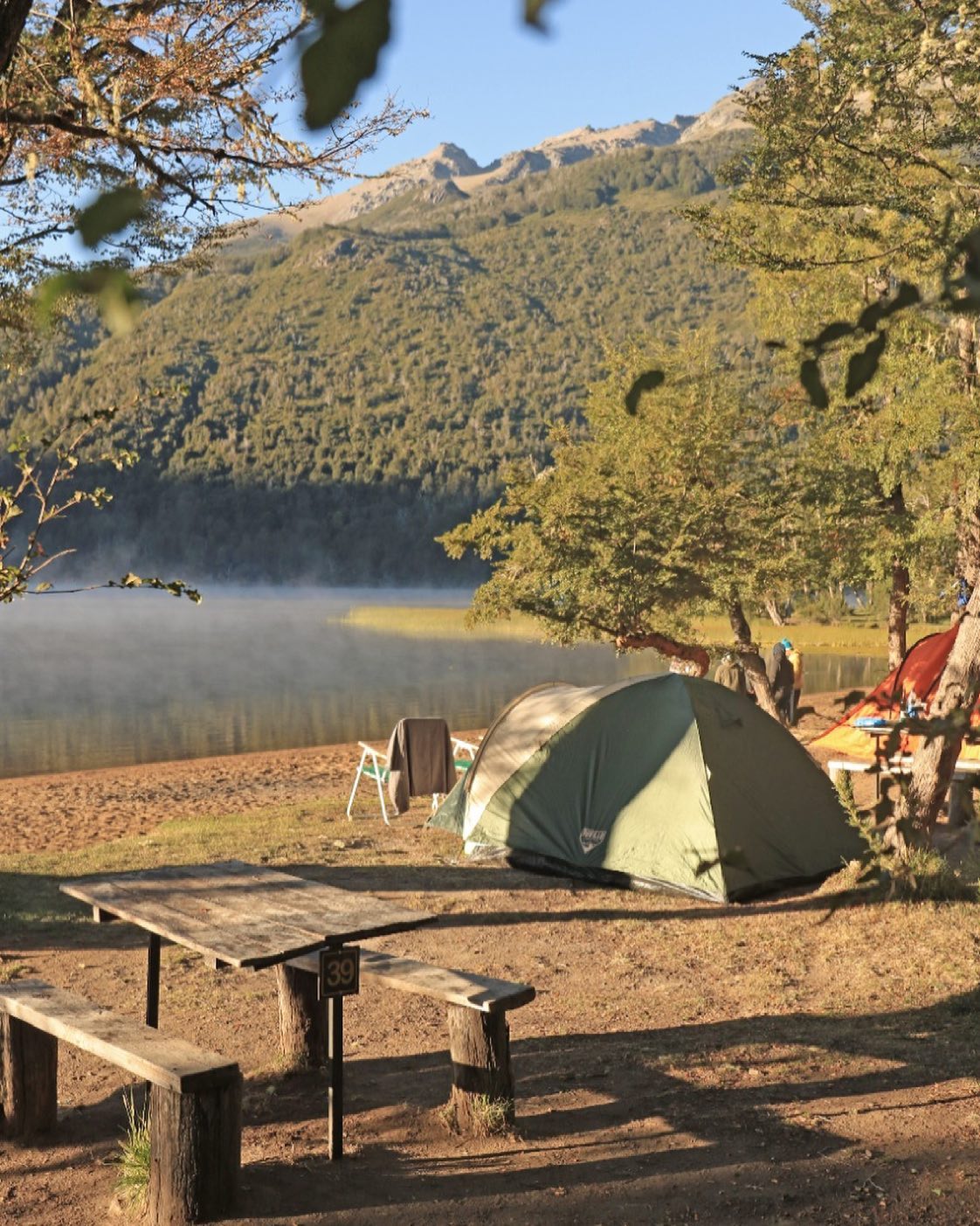 La imagen muestra una escena de campamento en un entorno natural frente al lago que se extiende horizontalmente.
En el centro de la imagen, se ve una carpa y una mesa de picnic
El área de campamento está rodeada de árboles y vegetación con tonos verdes y marrones.