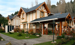 La imagen muestra una serie de casas adosadas de estilo alpino, ubicadas en un entorno natural con árboles. Las casas tienen un diseño distintivo con paredes de color claro y detalles de madera, y techos a dos aguas pronunciados. 