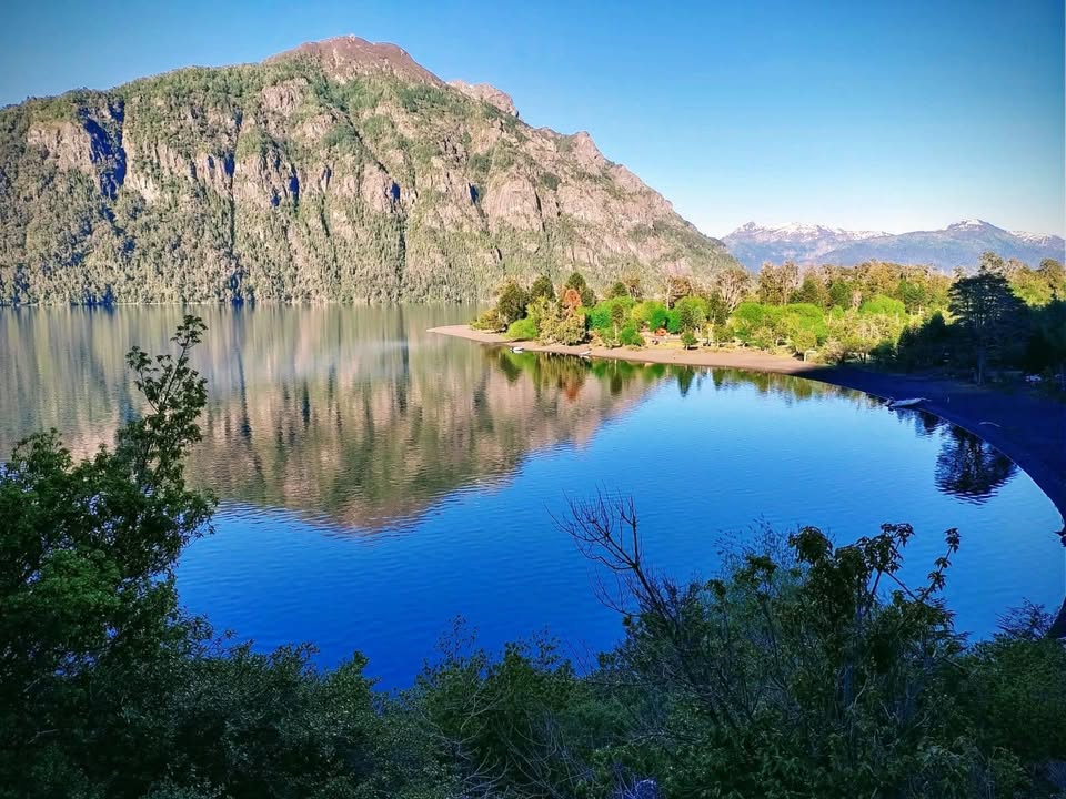 Imagen de un gran lago donde se refleja la montaña que est´+a detrás con mucha vegetación, bordeando el lago una playa de arena volcánica negra y bosque nativo.