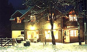 En la imagen se observa un edificio de dos pisos, con ventanas iluminadas que irradian una luz cálida. El entorno está cubierto de nieve, incluyendo el suelo, los árboles sin hojas y el techo a dos aguas de la casa, lo que indica que es de noche y hace frío. Hay algo de mobiliario exterior cubierto de nieve.
A pesar del frío exterior y la oscuridad de la noche invernal, la luz que emana de las ventanas sugiere un interior cálido y confortable, invitando a resguardarse. Puede evocar una sensación de hogar, seguridad y la magia de una noche de invierno.