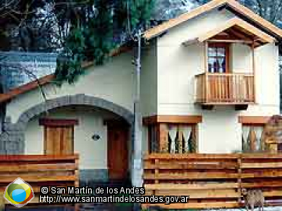 La imagen muestra una casa de dos pisos con un diseño arquitectónico distintivo, ubicada en un entorno natural. La casa tiene un techo a dos aguas pronunciado y está construida con una combinación de madera y piedra. Al ver la imagen, se siente una sensación de calma, calidez y simplicidad acogedora.