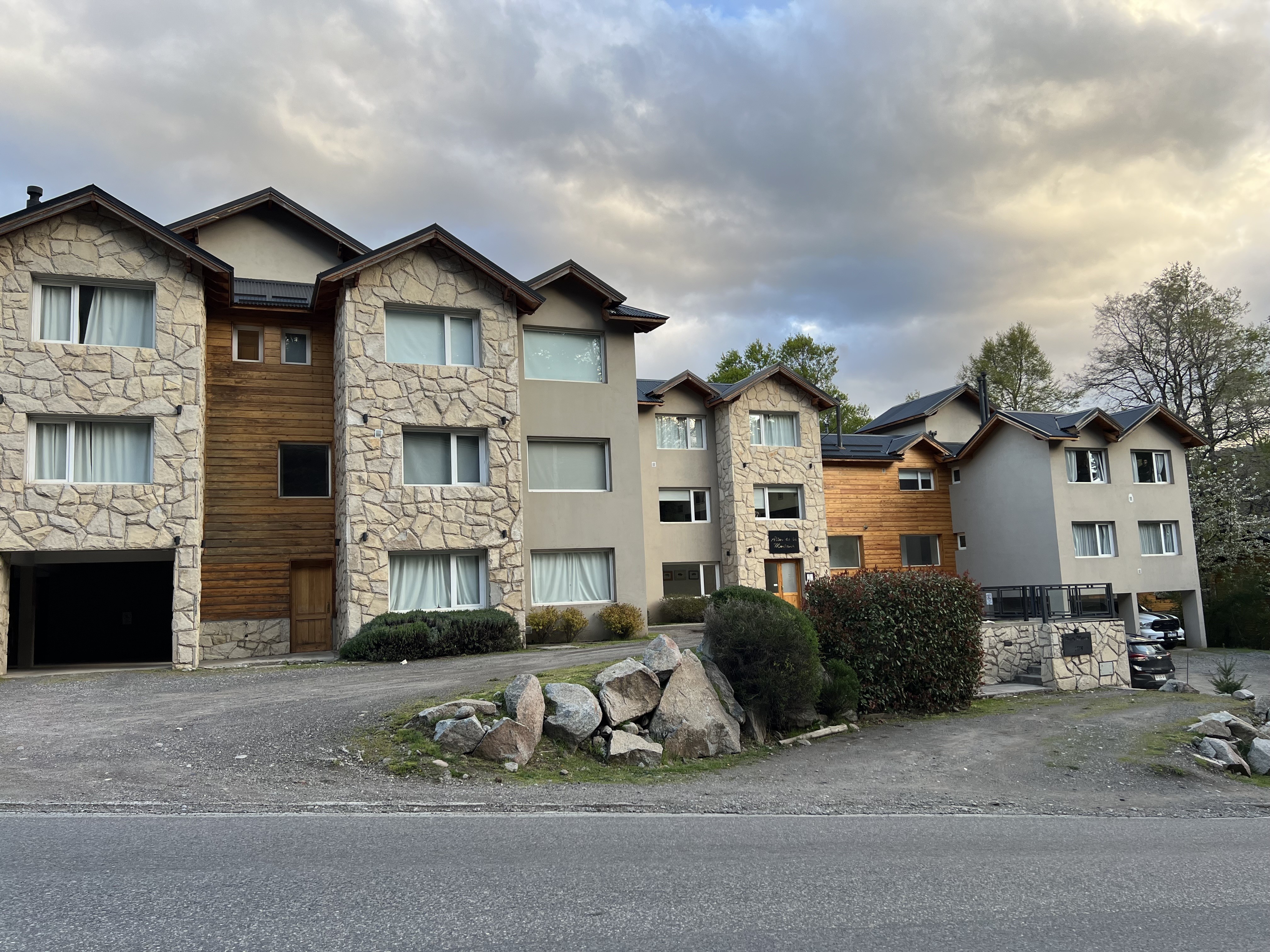 La imagen muestra un conjunto de edificios residenciales de estilo moderno-rústico. Las fachadas combinan piedra clara y revestimiento de madera, con techos inclinados oscuros. Departamentos  de tres pisos.

Al frente hay un camino de tierra que funciona como acceso y estacionamiento, junto a una calle asfaltada en primer plano. Se ven algunas rocas grandes decorativas y arbustos bien cuidados cerca de la entrada. A la derecha hay más unidades con balcones y un auto estacionado parcialmente visible.


