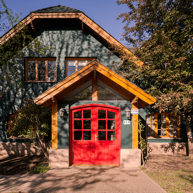 En la imagen, se observa la fachada de una casa de estilo rústico, que se integra bien con el entorno natural de San Martín de los Andes. La casa está construida predominantemente con madera, pintada de un color verde oscuro y una puerta de dos hojas en color rojo, lo que le da un aspecto distintivo. El techo es a dos aguas y hay un porche de entrada con un alero de madera clara que contrasta con el color de la casa. Al ver la imagen, se siente una sensación de encanto, calidez y originalidad