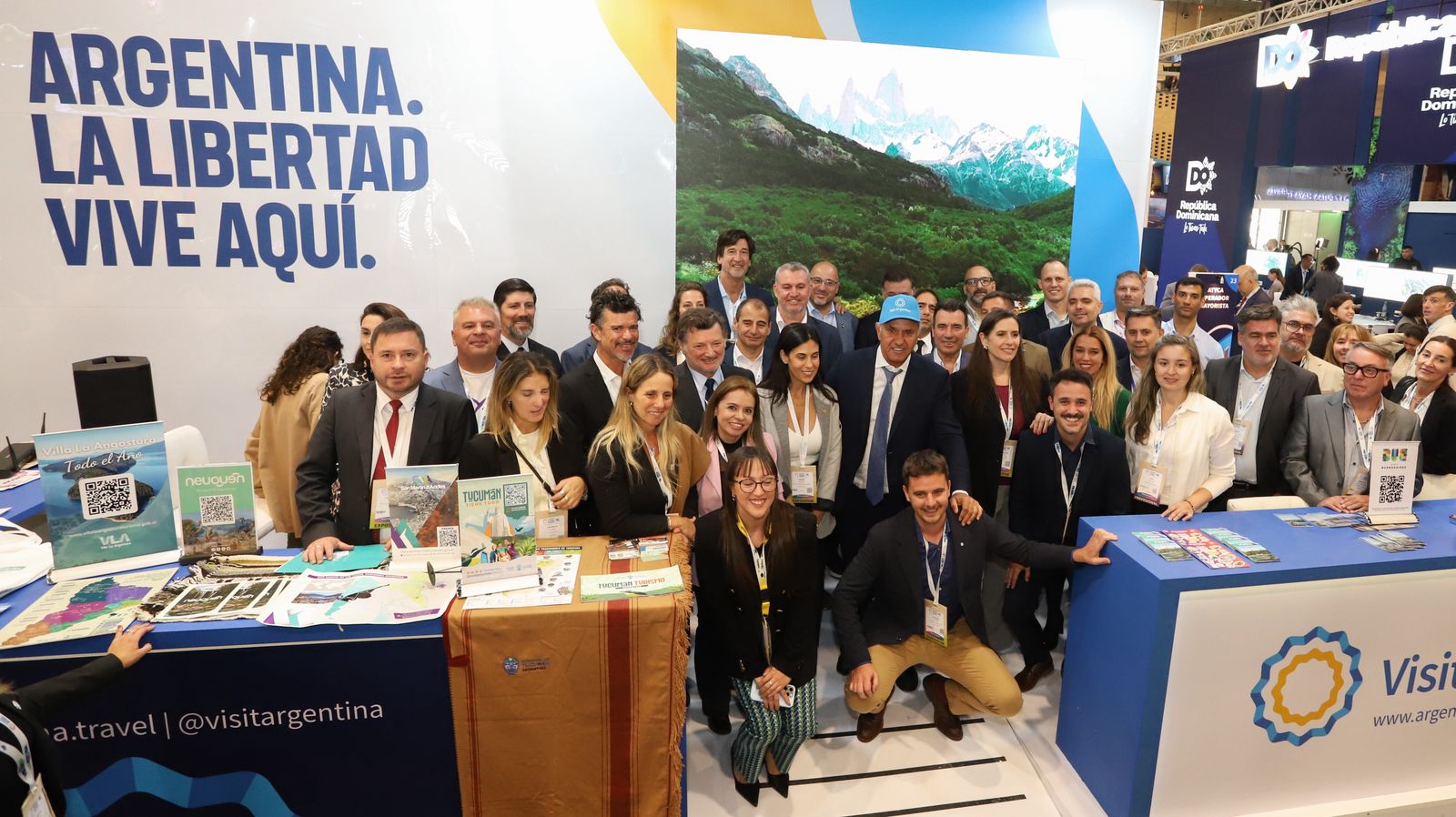 La imagen muestra un grupo numeroso de personas reunidas en  un evento institucional. Están posando para una foto grupal detrás de una mesa o mostrador con material promocional.
En el fondo se ve un banner grande con un paisaje natural (montañas y lago) y un texto que dice “Argentina, la ruta natural”, lo que sugiere que se trata de un evento relacionado con turismo o promoción del país. Varias personas levantan los brazos en gesto de celebración o entusiasmo.
La mayoría viste ropa formal o se