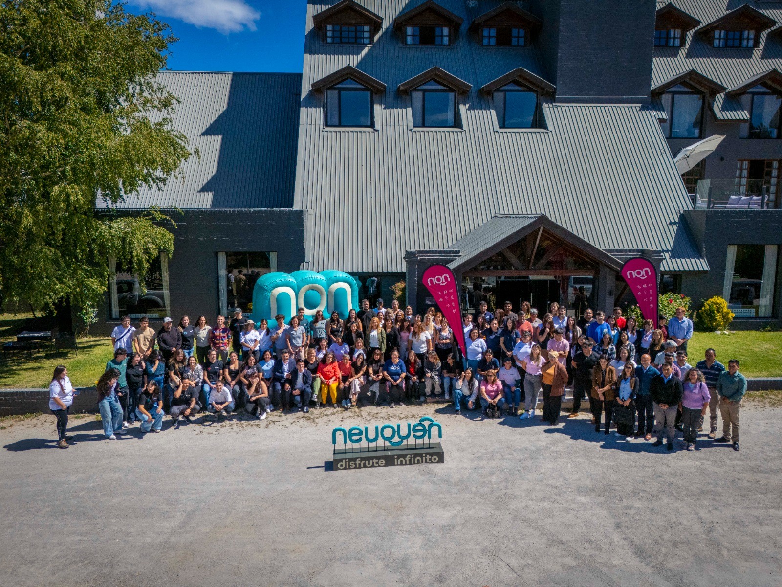 En la imagen se observa un grupo grande de gente posando para un foto, detrás de ellos un edificio con techo de chapas. 