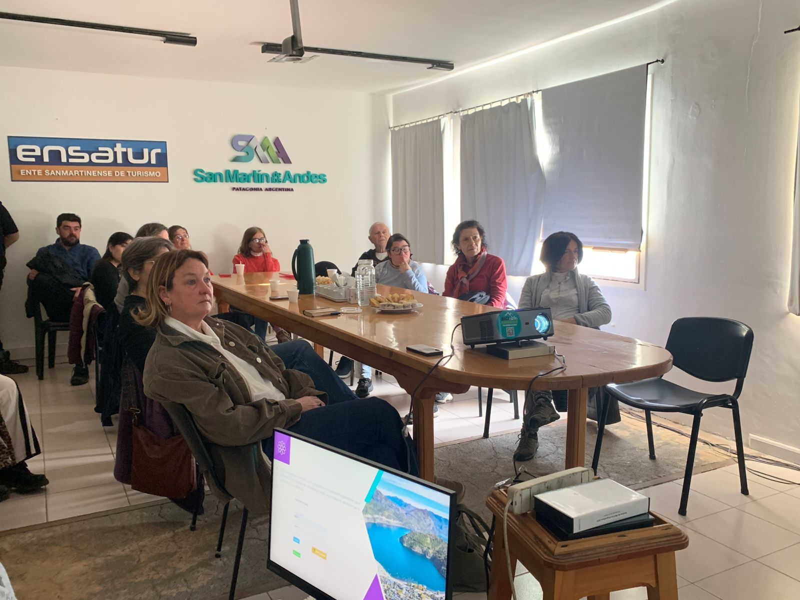La imagen muestra un grupo de personas sentadas alrededor de una mesa larga en un salón luminoso.
