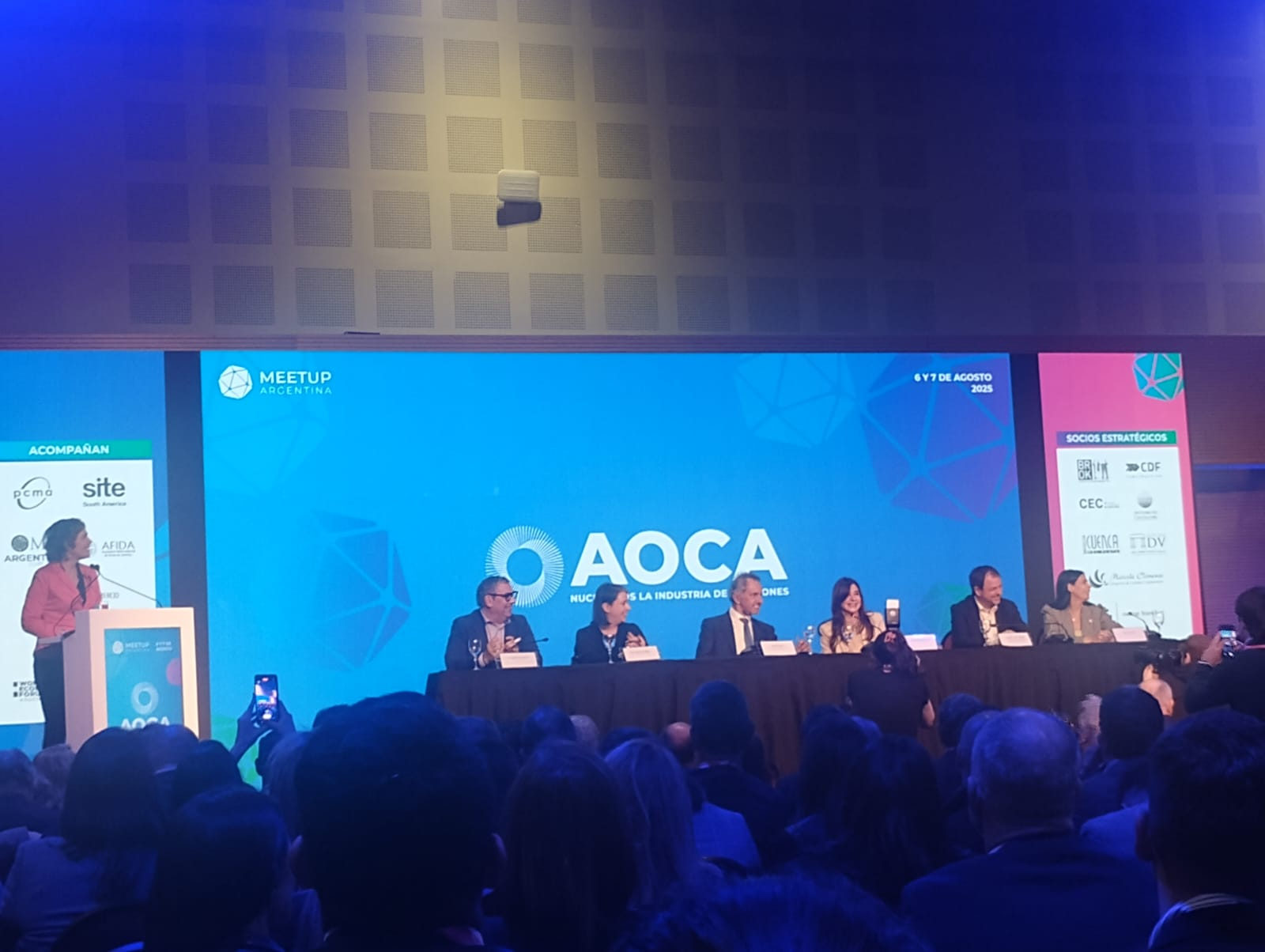 En la foto se puede ver gente sentada con un pantalla detrás que muestra el evento.